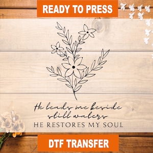 May include: A black floral design with the text "He leads me beside still waters. He restores my soul." The words "READY TO PRESS" and "DTF TRANSFER" are in orange boxes.