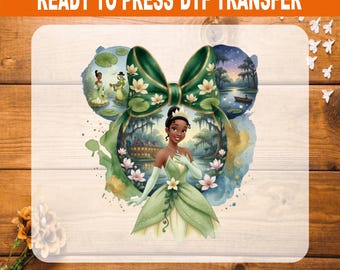 May include: A Ready to Press DTF transfer featuring a central illustration of a woman in a green gown with a large green bow above her head. The design includes a bayou scene with a house, lily pads, and a small boat, all in watercolor style.