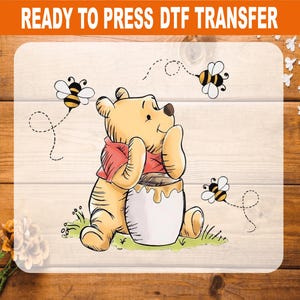 May include: A DTF transfer sheet featuring a cartoon yellow bear wearing a red shirt, sitting and leaning on a white honey pot. Three small bees with black and yellow stripes fly around the bear. The text READY TO PRESS DTF TRANSFER is at the top.