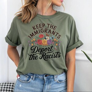 May include: An olive green t-shirt with rolled sleeves, featuring the text "Keep the Immigrants, Deport the Racists" in black script. The text is above a floral design with red, yellow, and purple flowers, and butterflies.