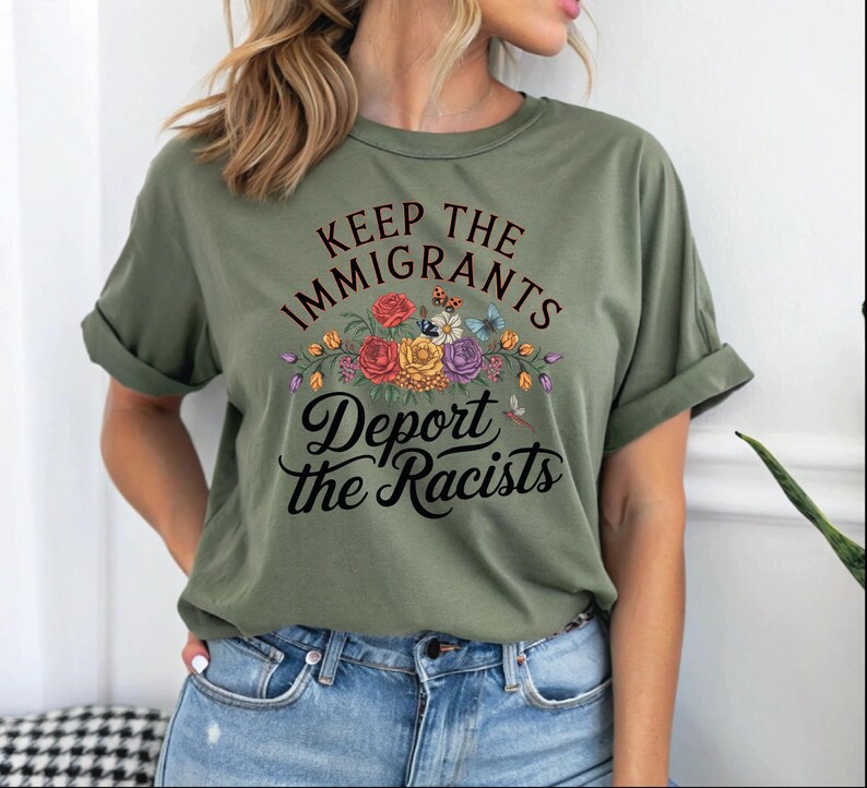 May include: An olive green t-shirt with rolled sleeves, featuring the text "Keep the Immigrants, Deport the Racists" in black script. The text is above a floral design with red, yellow, and purple flowers, and butterflies.