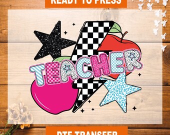 May include: A colorful graphic design with the word "TEACHER" in a bold, patterned font. The design includes a checkered lightning bolt, a pink apple, and a black star. The design is ready to press onto a t-shirt or other fabric.