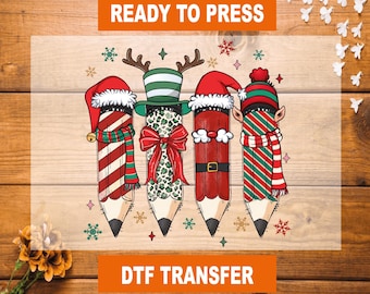 May include: A DTF transfer featuring four pencils dressed in festive holiday attire. Each pencil is decorated with a Santa hat, elf ears, or reindeer antlers, and a matching scarf. The words "READY TO PRESS" and "DTF TRANSFER" are displayed in orange.