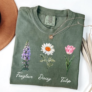 May include: A green t-shirt with a foxglove, daisy, and tulip design. The flowers are illustrated in watercolor style. The text "Foxglove", "Daisy", and "Tulip" are printed below the flowers.