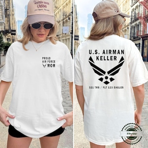 May include: A white t-shirt featuring black text and graphics. The front displays 'PROUD AIR FORCE MOM' with an emblem. The back features 'U.S. AIRMAN KELLER' above a large Air Force logo and the text '321 TRS / FLT 123 EAGLES' in black ink.