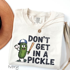 May include: Ivory-colored t-shirt featuring a cartoon pickle wearing a baseball cap and holding a bat and ball. The shirt has the phrase "DON'T GET IN A PICKLE" printed in black. A gold necklace and a brown hat are also visible.