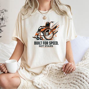 May include: An ivory short-sleeve t-shirt featuring a graphic of a wheelchair with orange and black motion lines. Below the graphic, the text reads "BUILT FOR SPEED, NOT STAIRS" in black capital letters with an orange line underneath the word STAIRS.