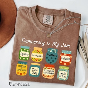 May include: Espresso-colored t-shirt with the text "Democracy Is My Jam" and illustrations of jars labeled with words like "Equality" and "Social Justice." A silver necklace with a pendant is draped on the shirt.