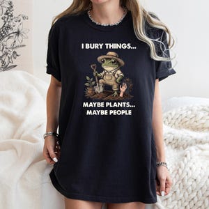 May include: Black t-shirt with a humorous graphic of a frog in overalls and a hat holding a shovel. The text reads "I bury things... Maybe plants... Maybe people."