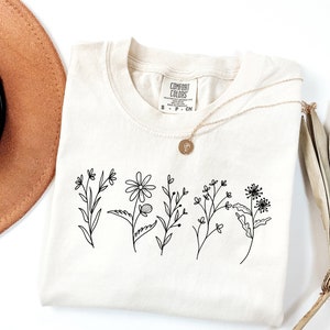 K&ouml;nnte beinhalten: Ein cremefarbenes T-Shirt mit einer schwarzen Linienzeichnung von Wildblumen. Das Shirt hat ein Etikett mit der Aufschrift "Comfort Colors" und den Gr&ouml;&szlig;en S, P und CN.