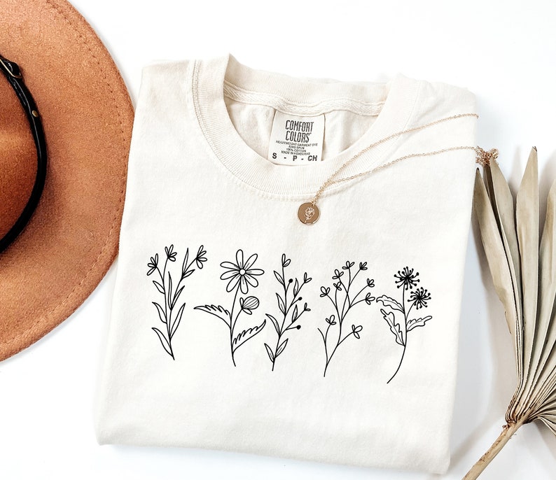K&ouml;nnte beinhalten: Ein cremefarbenes T-Shirt mit einer schwarzen Linienzeichnung von Wildblumen. Das Shirt hat ein Etikett mit der Aufschrift "Comfort Colors" und den Gr&ouml;&szlig;en S, P und CN.
