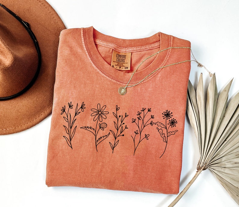 K&ouml;nnte beinhalten: Ein korallenfarbenes T-Shirt mit einer einfachen schwarzen Linienzeichnung von f&uuml;nf verschiedenen Blumen. Das Shirt ist gefaltet und liegt flach auf einer wei&szlig;en Oberfl&auml;che.