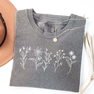 K&ouml;nnte beinhalten: Ein graues T-Shirt mit einem wei&szlig;en gestickten Design aus f&uuml;nf verschiedenen Blumen. Das Shirt hat einen Rundhalsausschnitt und ist aus einem weichen, bequemen Stoff gefertigt.
