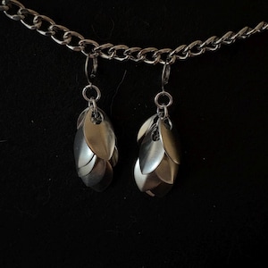 May include: A pair of silver-toned scale earrings suspended from a matching silver-toned metal chain. Each earring consists of multiple overlapping metallic scales arranged in a teardrop shape, attached to a circular jump ring and a lever-back clasp.
