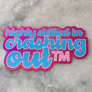 May include: A pink and blue sticker with the text "highly skilled in crashing out TM". The sticker has a glittery finish and a rounded rectangular shape. The text is in a playful, cursive font.
