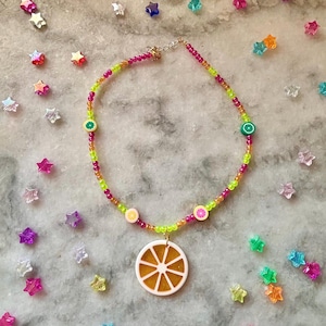 Op de afbeelding: Een kleurrijke kralenketting met een citrusvruchten thema. De ketting heeft een mix van roze, groene en oranje kralen, met fruit slice bedels en een grote sinaasappelschijf hanger. De ketting is geplaatst tegen een achtergrond van stervormige confetti.