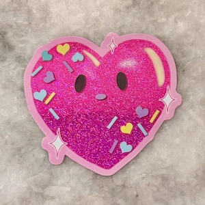 May include: A pink heart-shaped sticker with a glittery texture and a cartoon face. The sticker is decorated with colorful sprinkles, including hearts and rectangles. White star accents are on the edges.