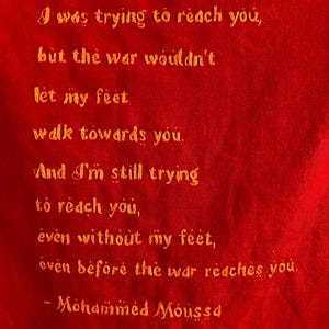 May include: A vibrant red fabric featuring yellow embroidered text that reads: I was trying to reach you, but the war wouldn't let my feet walk towards you. And I'm still trying to reach you, even without my feet, even before the war reaches you. - Mohammed Moussa