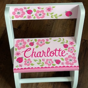 May include: A white wooden step stool with a pink floral design and the name "Charlotte" in cursive. The design includes pink flowers, green leaves, and ladybugs. The stool is painted with a white frame.