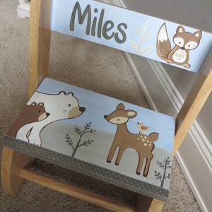May include: Wooden child's chair with a light blue back and seat. The back of the chair has the name "Miles" in brown lettering, a fox, and gold-colored leaves. The seat features a bear, deer, and trees.