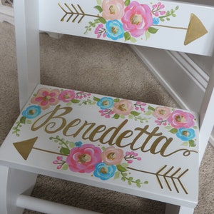May include: White wooden step stool with floral and arrow designs. The step stool features pink, blue, and peach flowers with gold arrow accents. The name "Benedetta" is written in gold script on the step.