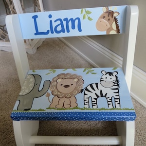 May include: A white wooden step stool with a light blue seat and backrest. The backrest has the name "Liam" in blue lettering and a giraffe illustration. The seat features an elephant, lion, and zebra design.