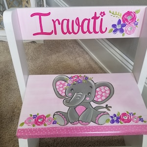 May include: A pink and white step stool with the name "Irawati" in pink script. The stool features a painted gray elephant with a floral crown and pink polka dot ears. Floral accents in pink and purple.