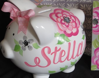 May include: White ceramic piggy bank with a pink ribbon, pink and gray flowers, and the name "Stella" painted on the side.