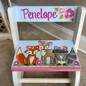 May include: A white wooden step stool with a light blue seat and backrest. The backrest has the name "Penelope" in pink script and pink floral accents. The seat features a fox, deer, and raccoon with floral crowns.
