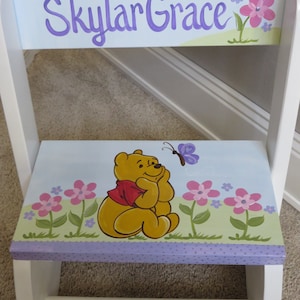 May include: A white wooden step stool with a painted design featuring Winnie the Pooh, flowers, and the name "Skylar Grace" in purple script. The stool has a light blue background and a purple polka-dot border.
