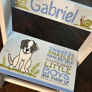 May include: A white wooden step stool with a blue and white painted design. The stool features a cartoon dog, green leaves, and the text "Gabriel, Snakes, Snails, Puppy Dog Tails, That's What Little Boys Are Made Of".