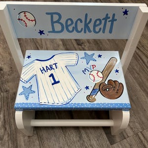 May include: A light blue and white wooden step stool featuring a baseball theme. The backrest displays the name Beckett and a baseball. The seat shows a white jersey with HART 1, a baseball bat, a brown glove, a baseball, and blue stars.
