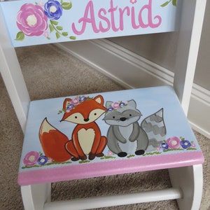 May include: A hand-painted wooden child's chair with a light blue seat and back. The seat features a fox and raccoon with floral crowns. The backrest displays the name "Astrid" in pink script, surrounded by flowers. The chair is painted white.