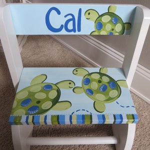 May include: A white wooden step stool with a light blue seat and backrest. The seat and backrest feature green turtles with blue and green spots. The backrest has the name "Cal" in blue lettering.