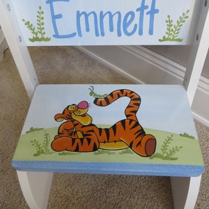 May include: A white wooden step stool with a blue and green painted design featuring a cartoon tiger and the name "Emmett".