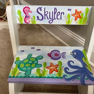 May include: A white wooden step stool with a hand-painted ocean theme. The design includes a seahorse, starfish, turtle, fish, and octopus. The name "Skyler" is written in purple script on the top step.