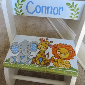 May include: A white wooden step stool with a blue and green painted top. The top features a green leafy design, the name "Connor" in blue lettering, and a painted scene of an elephant, giraffe, and lion.