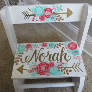 May include: White wooden step stool with a floral design in pink, teal, and gold. The name "Norah" is written in gold script on the step. Arrows and floral accents decorate the back and step.