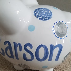 May include: White ceramic piggy bank with blue and gray polka dots and a wavy design. The name "Carson" is written in blue on the side.