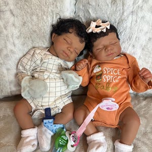 May include: Two realistic baby dolls are lying down. One wears a white and beige checkered onesie, the other an orange onesie with "Spice Baby" text. Both have dark hair, closed eyes, and pacifiers.