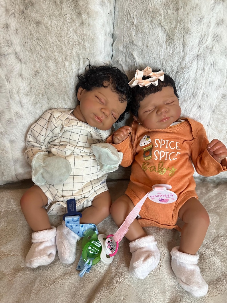May include: Two realistic baby dolls are lying down. One wears a white and beige checkered onesie, the other an orange onesie with "Spice Baby" text. Both have dark hair, closed eyes, and pacifiers.