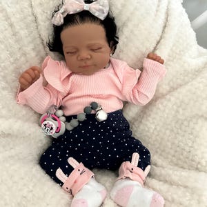 May include: A realistic baby doll dressed in a pink ruffled top, navy blue polka dot trousers, and bunny-themed socks. The doll has a pink bow and a pacifier with a Minnie Mouse design.