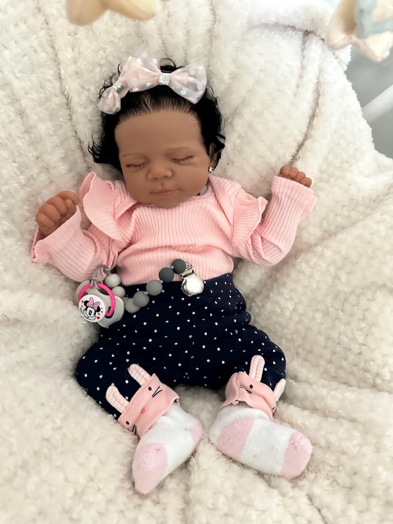 May include: A realistic baby doll dressed in a pink ruffled top, navy blue polka dot trousers, and bunny-themed socks. The doll has a pink bow and a pacifier with a Minnie Mouse design.