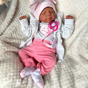 May include: A realistic baby doll dressed in a pink polka-dot bow hat, a grey zip-up jacket, a striped shirt with a heart graphic, and pink trousers. The doll has a pink dummy and is lying on a fluffy white blanket.