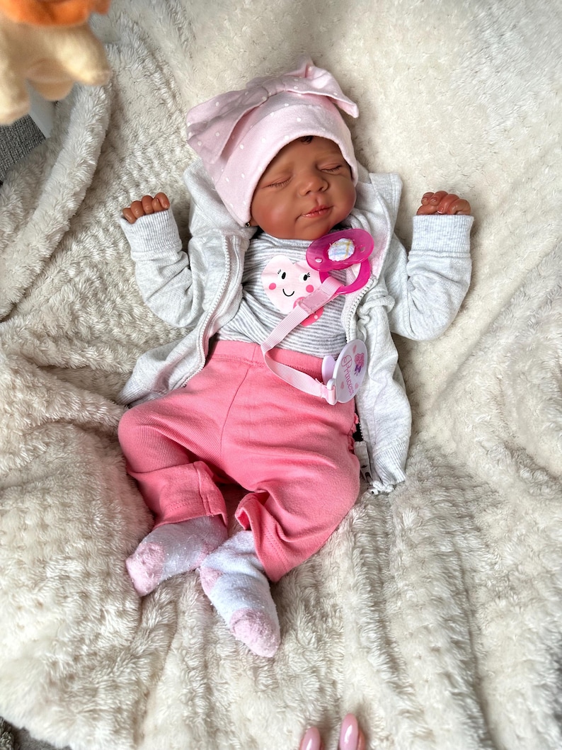 May include: A realistic baby doll dressed in a pink polka-dot bow hat, a grey zip-up jacket, a striped shirt with a heart graphic, and pink trousers. The doll has a pink dummy and is lying on a fluffy white blanket.