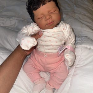 May include: A realistic baby doll with dark skin and closed eyes, wearing a white onesie with a floral pattern, pink trousers, and white socks. The doll has dark, curly hair and is lying on a white surface.