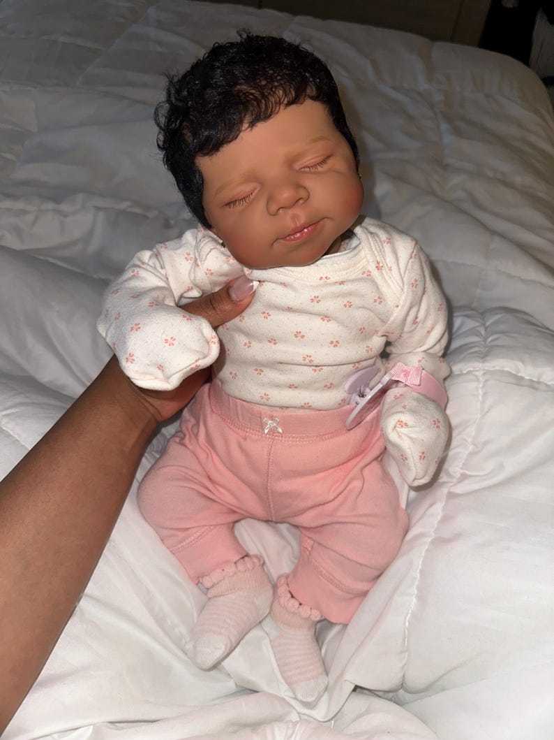 May include: A realistic baby doll with dark skin and closed eyes, wearing a white onesie with a floral pattern, pink trousers, and white socks. The doll has dark, curly hair and is lying on a white surface.