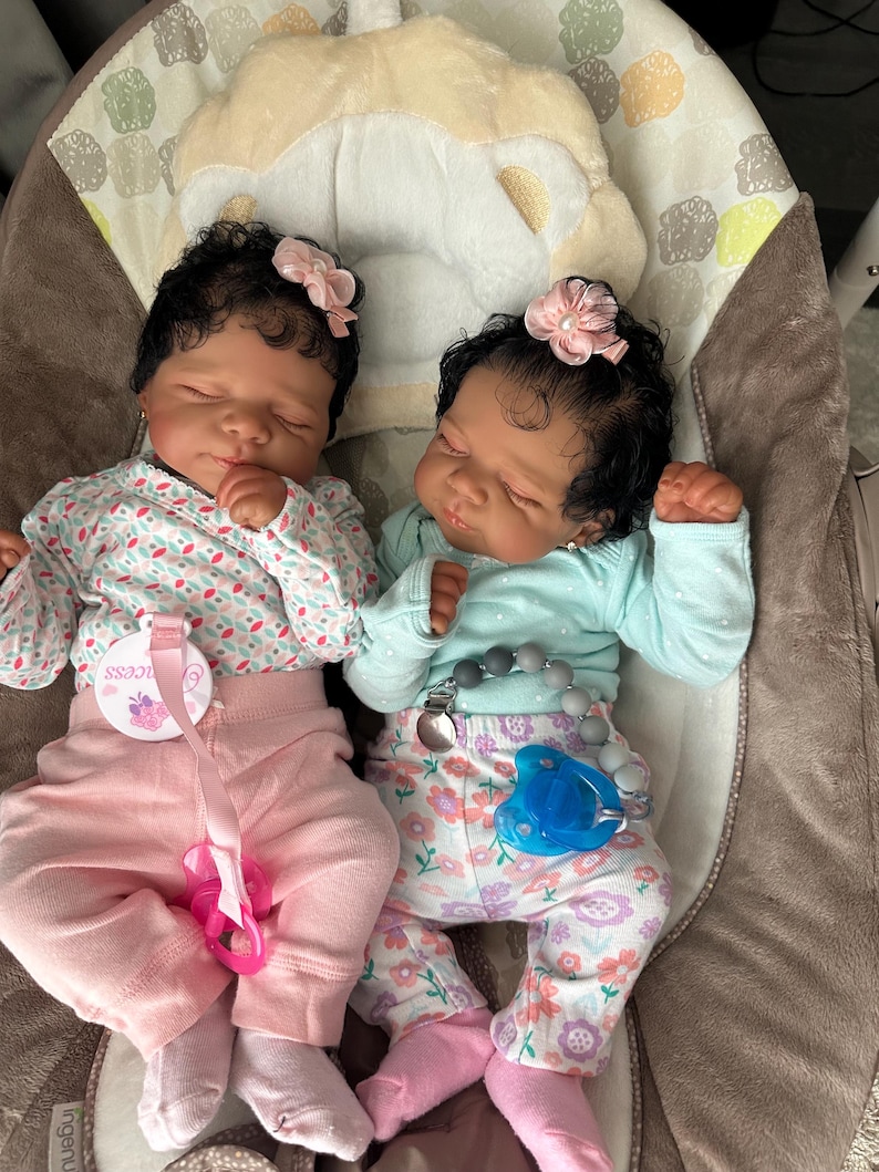 May include: Two realistic baby dolls with dark skin and curly black hair, each wearing a pink bow. One doll is in pink trousers and a floral top, the other in floral trousers and a light blue top. Both have dummies.