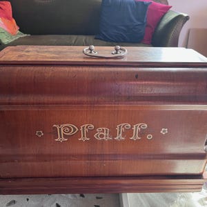 Può includere: Una custodia per macchina da cucire vintage in legno con una maniglia in metallo e la scritta "Pfaff" incisa sul davanti.