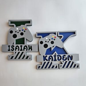 May include: Two personalized wooden game controller-shaped name signs. One sign features the name "ISAIAH" in black, with a gray controller and green accents. The other sign has the name "KAIDEN" in black, with a gray controller and blue accents.